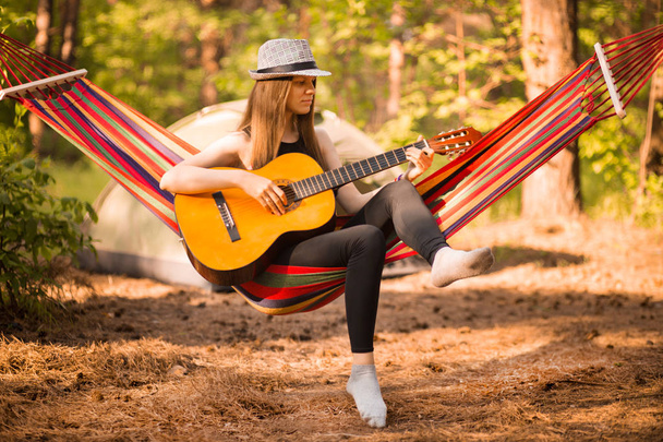 Vrouw met hoed spelen gitaar en ontspannen in hangmat opknoping tussen pijnbomen op de achtergrond. Camping rustconcept - Foto, afbeelding