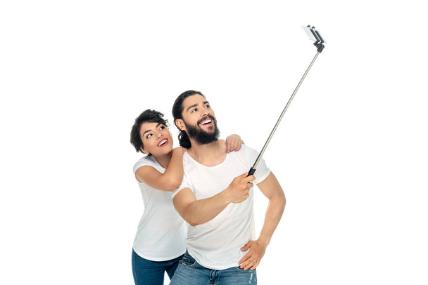 χαρούμενο Latin άνθρωπος χαμογελά κοντά μελαχρινή γυναίκα, ενώ κρατώντας selfie ραβδί και παίρνοντας selfie απομονώνονται σε λευκό  - Φωτογραφία, εικόνα