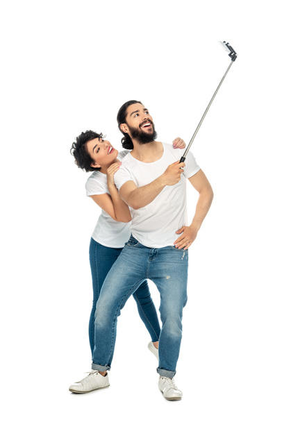 szczęśliwy łaciński człowiek uśmiechnięty w pobliżu brunetka kobieta trzymając selfie kij i biorąc selfie na białym tle  - Zdjęcie, obraz