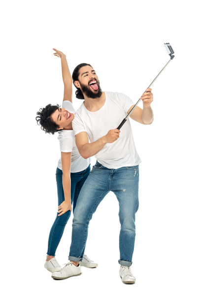 ευτυχισμένος Λάτιν άνθρωπος δείχνει τη γλώσσα κοντά μελαχρινή γυναίκα με τεντωμένα χέρια, κρατώντας selfie ραβδί και παίρνοντας selfie απομονώνονται σε λευκό  - Φωτογραφία, εικόνα