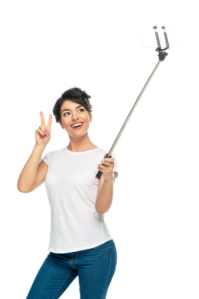 happy latin woman showing peace sign while holding selfie stick and taking selfie isolated on white  - Φωτογραφία, εικόνα