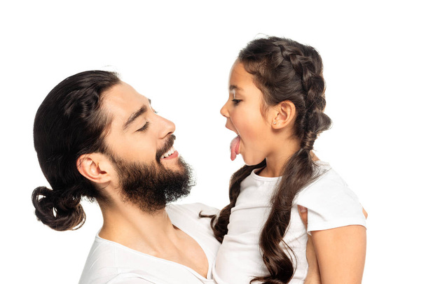 szczęśliwy łaciński ojciec uśmiechnięty patrząc na cute córki pokazując język na białym tle  - Zdjęcie, obraz