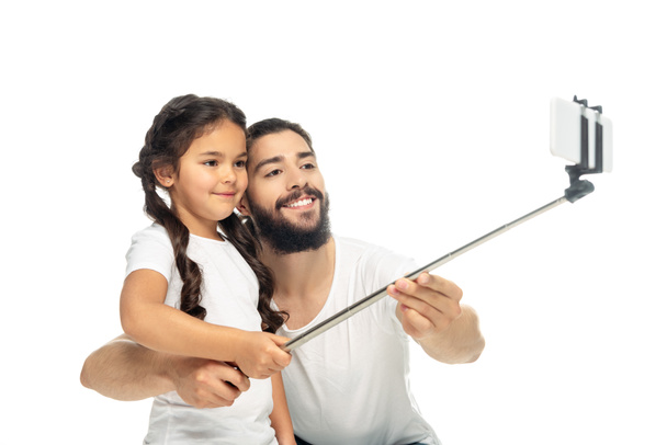 ευτυχισμένο παιδί κρατώντας selfie ραβδί, ενώ παίρνοντας selfie με το λατινικό πατέρα απομονώνεται σε λευκό  - Φωτογραφία, εικόνα