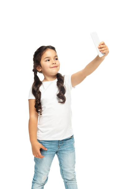 白で隔離されたスマートフォンで自分撮りを取る幸せなラテンの子供  - 写真・画像