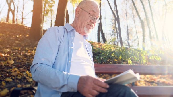 Senior man with a beard and wearing glasses reading a book in the park. - Фото, изображение