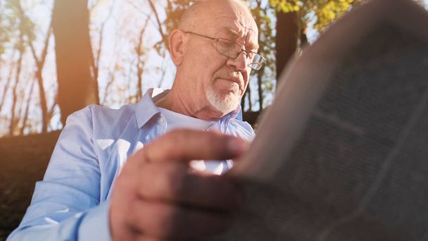 Old man relaxing at park. Senior man with a beard and wearing glasses reading newspaper in the park. - Photo, Image