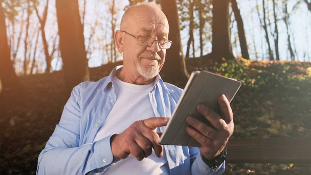 Active senior man walking in the park and using a tablet sitting in the park on the bench. - Fotoğraf, Görsel