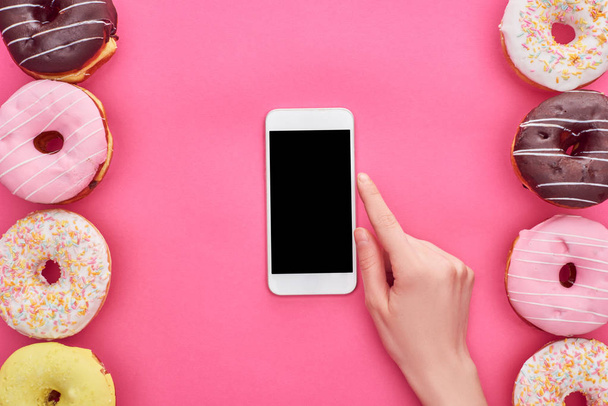 cropped view of woman pointing with finger at smartphone on pink background with tasty doughnuts - Foto, Bild
