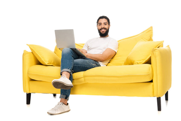 ευτυχισμένος μουσάτος Λάτιν άνθρωπος χρησιμοποιώντας φορητό υπολογιστή, ενώ κάθεται σε κίτρινο καναπέ απομονωθεί σε λευκό  - Φωτογραφία, εικόνα