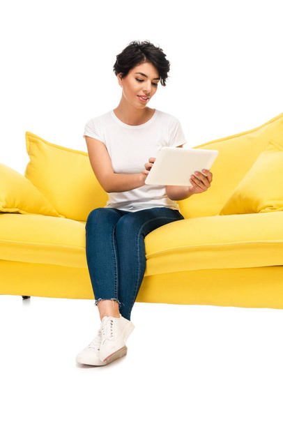 happy latin woman using digital tablet while sitting on sofa isolated on white  - Zdjęcie, obraz