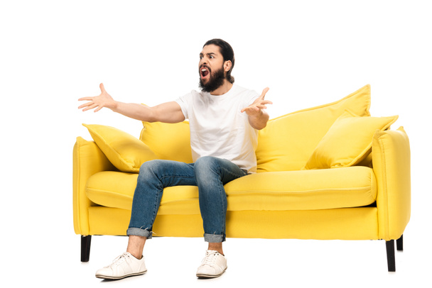 бородатый латинос смотрит чемпионат, сидя на диване и жестикулируя изолированным на белом
  - Фото, изображение