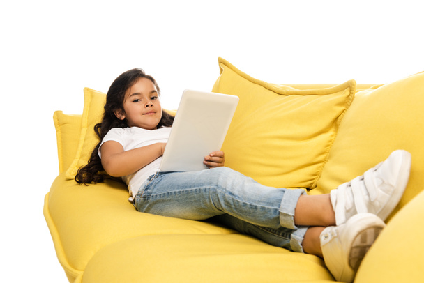 selektywne skupienie szczęśliwego łacińskiego dzieciaka za pomocą cyfrowego tabletu leżącego na kanapie na białym tle  - Zdjęcie, obraz