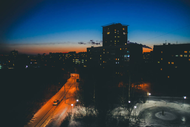 Εκπληκτικές σιλουέτες με φωτεινά παράθυρα στο φόντο του θερμού ουρανού του ηλιοβασιλέματος. Καπνός από σωλήνα πάνω από την πόλη την αυγή. Λαμπερά παράθυρα στο λυκόφως. Μινιμαλιστικό αστικό τοπίο. Υπέροχο αστικό Ανατολή. - Φωτογραφία, εικόνα