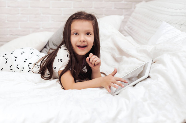 little child girl lies in bed uses digital tablet. - 写真・画像