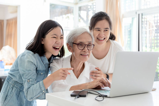 Anne ve kızı internette sörf;büyükanne ile ilginç bir şey izlerken,mutlu gülümseyen asyalı yaşlı kadın ise kızı ve torunu evde masada dizüstü bilgisayar kullanarak, konsept aile, teknoloji  - Fotoğraf, Görsel