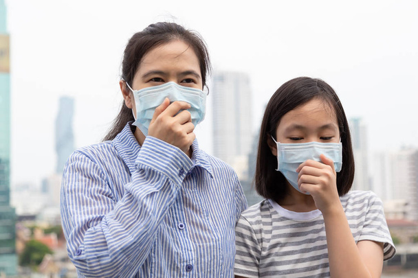アジアの女性や母と娘は、フェイスマスク保護、背景として市の建物の大気汚染のためにフェイスマスクを着用しているかわいい子供と大人の女性、医療マスクを持つ病気の女の子;汚染、粉塵アレルギーの概念で咳に苦しんでいます  - 写真・画像