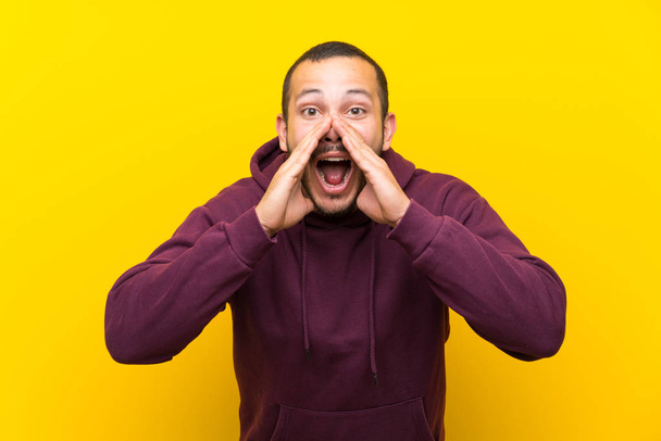 Colombian man with sweatshirt over yellow wall shouting and announcing something - Photo, Image