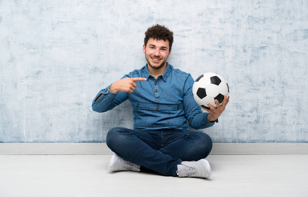 Νέος άνθρωπος κάθεται στο πάτωμα κρατώντας μια μπάλα ποδοσφαίρου - Φωτογραφία, εικόνα