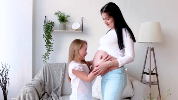 Pregnant woman and her little daughter having fun indoors. Maternity. Daughter kissing belly of pregnant mother  - Záběry, video