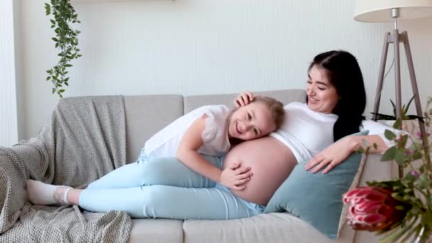 Pregnant woman and her little daughter having fun indoors. Maternity. Young mother on sofa - Footage, Video