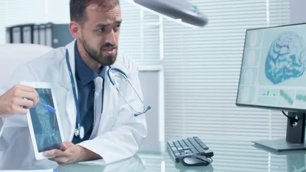 Médico en la oficina de moder con escritorio de cristal hablando y apuntando a una pantalla de tableta digital con una radiografía en él
 - Metraje, vídeo