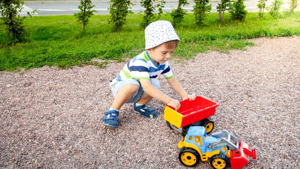 Portrait of cute 3 years old toddler boy sitting on the playground at park and playing with colorful plastic toy truck. Child having fun and playing outdoors with toys - Foto, Imagen
