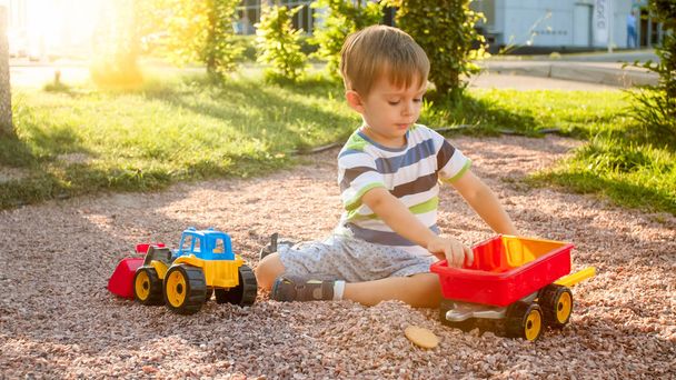 Nahaufnahme Bild von niedlichen kleinen Jungen, die auf dem Palyground mit Spielzeug spielen. Kinder haben Spaß mit LKW, Bagger und Anhänger. er gibt sich als Bauunternehmer oder Fahrer aus - Foto, Bild