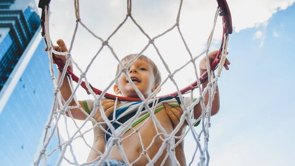 Close-up imagem de bonito sorrindo criança menino segurando e pendurado no anel de basquete em sportrs playground na cidade
 - Foto, Imagem
