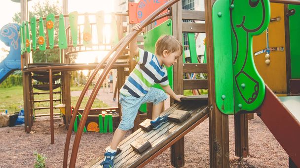 Photo of adorable toddler boy climbing and crawling on wooden staircase on children palyground at park - 写真・画像