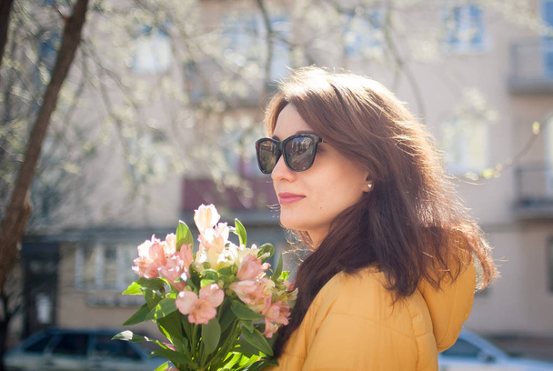Outdoors portrait of stylish attractive brunette woman in sunglasses walking outdoors with big bouquet of colorful flowers during sunny spring day while the sun is shining - Foto, immagini