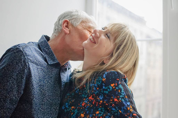 Femme d'âge moyen chaude et sexy profitant d'embrasser son mari âgé debout près de la fenêtre ouverte à l'intérieur de leur maison. Couple avec différence d'âge
 - Photo, image