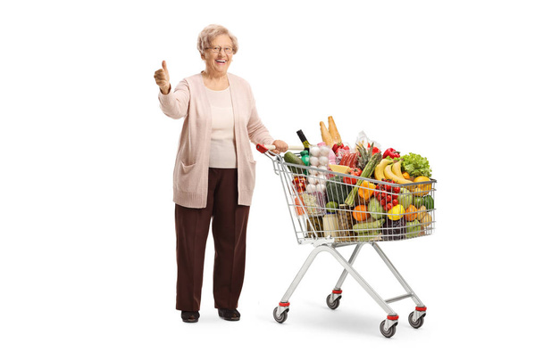 Full length portrait of a smiling senior woman posing with a shopping cart full of food and showing thumbs up isolated on white background - Фото, зображення