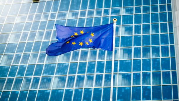 Образ флага ЕС, развевающегося на ветру против высокого офисного здания, выполненного из концерта и стекла. Концепция экомики, развития, правительства и политики
 - Фото, изображение