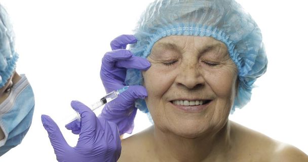 Пожилая женщина в защитной шляпе. Врач делает уколы лица пациенту
 - Фото, изображение