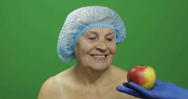 Ηλικιωμένη γυναίκα με προστατευτικό καπέλο. Ο γιατρός της δείχνει ένα μήλο. - Φωτογραφία, εικόνα
