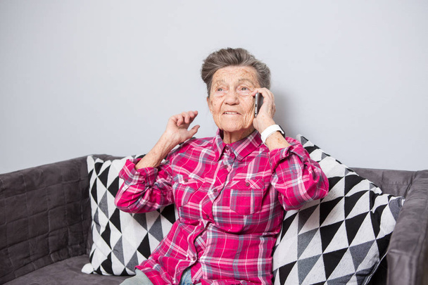 Theme old person uses technology. Mature happy joy smile active gray hair Caucasian wrinkles woman sitting home in living room on the sofa and using a mobile phone, calling and talking on the phone - Photo, Image