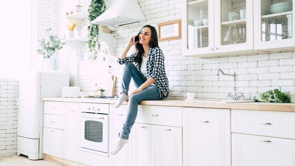 Beautiful cute smiling woman is using smart phone on the kitchen at home. - Fotó, kép