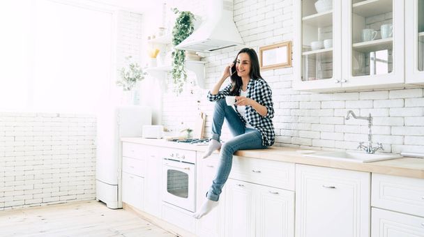 Beautiful cute smiling woman is using smart phone on the kitchen at home. - Photo, Image