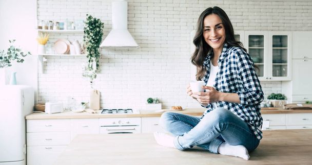 Cute lovely young woman in casual wear with coffee cup in hands is sitting on table in kitchen and relaxing. - Foto, Imagem