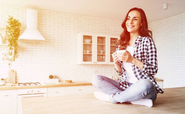 Cute lovely young woman in casual wear with coffee cup in hands is sitting on table in kitchen and relaxing. - Foto, Bild