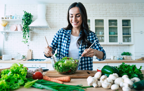 Bonito bonito jovem sorrindo mulher na cozinha está preparando uma salada vegan em roupas casuais
. - Foto, Imagem