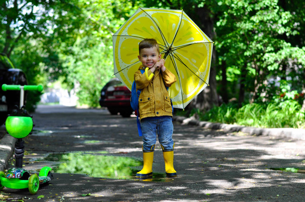  Ένα μικρό χαριτωμένο αγόρι σε ένα κίτρινο αδιάβροχο περπατάει στις λιμνούλες χαμογελώντας και κρατώντας μια ομπρέλα με ένα χρώμα λεμονιού. - Φωτογραφία, εικόνα