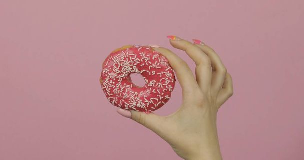 Жіноча рука тримає червоний, смачний, посипаний пончик на рожевому фоні
. - Фото, зображення