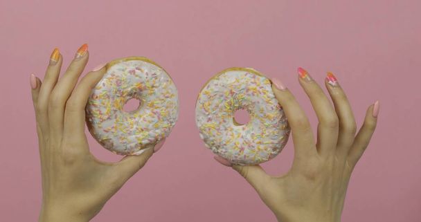 Жіночі руки тримають два білих, смачні, посипані пончики на рожевому фоні
. - Фото, зображення