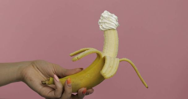 Woman hand holds banana with whipped cream on top of the fruit - Foto, imagen