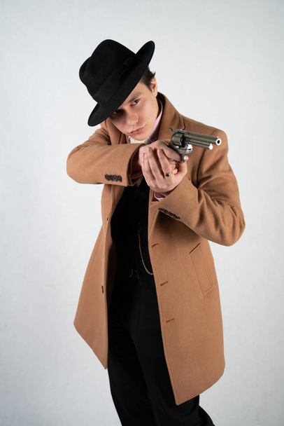 正式なスーツと帽子をかぶった若い男が、孤立したスタジオの白い背景に銃を手に - 写真・画像