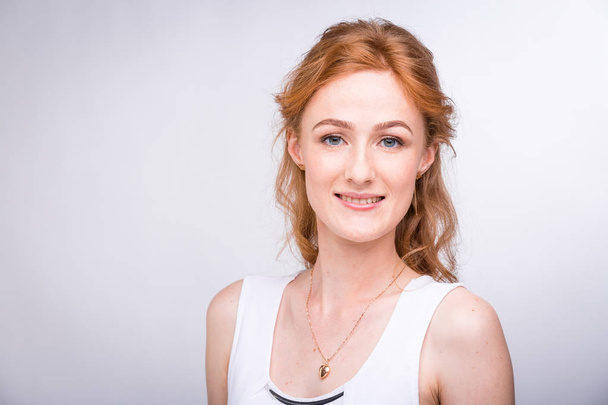 Portrait of a beautiful young female student with a smile in a white shirt of European, Caucasian nationality with long red hair and freckles on her face posing on a white background in the studio - Photo, Image
