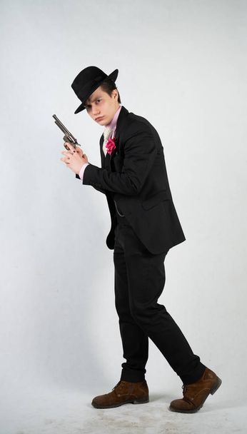 joven con un traje formal y un sombrero con una pistola en las manos sobre un fondo blanco solo en el estudio
 - Foto, Imagen