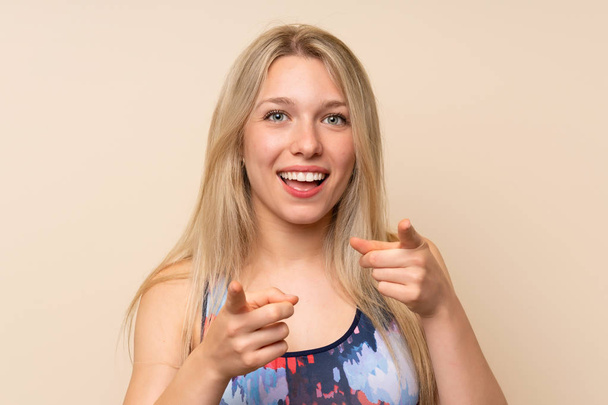 Молодая блондинка спортивная женщина над изолированной стеной указывает пальцем на тебя
 - Фото, изображение