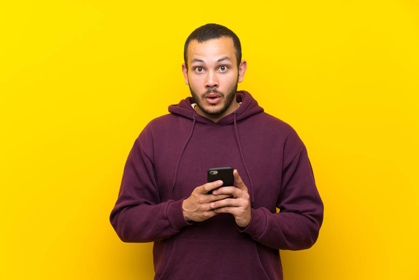 Colombian man with sweatshirt over yellow wall surprised and sending a message - Φωτογραφία, εικόνα
